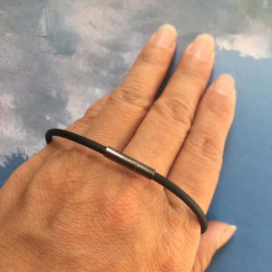 Op de afbeelding: Een zwarte rubberen armband met een zilveren metalen sluiting. De armband wordt om de pols gedragen.
