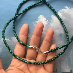 Può includere: Una collana di corda verde scuro con chiusura argentata. La collana è realizzata in cordoncino ritorto e ha un design semplice ed elegante. La chiusura è a moschettone standard.