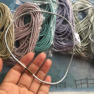 May include: A silver glittery cord necklace featuring a cylindrical brass-toned metal clasp. In the background, several coiled bundles of similar cords in gold, pink, teal, purple, and silver are individually packaged in clear plastic sleeves.