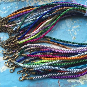 Puede incluir: Una colección de cordones trenzados de colores con cierres metálicos. Los cordones están dispuestos en forma de abanico y son de varios tonos de azul, verde, morado, rosa, naranja, amarillo y blanco.