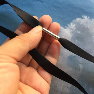 May include: A smooth black satin ribbon necklace featuring a cylindrical silver-toned metal magnetic clasp. The ribbon is thin and flat, connecting to the polished metal fastener which secures the two ends of the fabric band together.