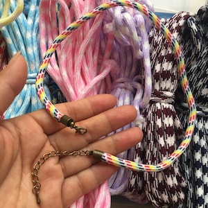 May include: A colorful rope necklace with a rainbow pattern, featuring a bronze-colored clasp and extender chain. The necklace is held in front of a backdrop of various rope bundles in pink, purple, and blue patterns.