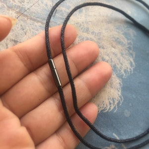 以下が含まれることがあります： 銀色の留め具が付いた黒い紐のネックレス。紐は二重になっており、手で持たれています。