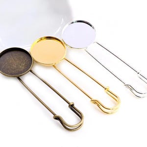 May include: Three different coloured metal safety pin brooches with round glass or metal cabochons. The cabochons are in gold, silver, and antique bronze.