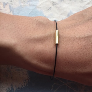 Könnte beinhalten: Ein schlichtes Armband aus schwarzem Band mit einem goldenen Balkenakzent.