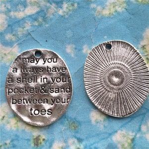 May include: Two silver metal charms. One charm has a sunburst design. The other charm has the text "may you always have a shell in your pocket & sand between your toes".