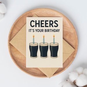 K&ouml;nnte beinhalten: Geburtstagskarte mit dem Text "CHEERS IT'S YOUR BIRTHDAY". Die Karte zeigt drei Cartoon-Biergl&auml;ser mit brennenden Kerzen. Die Karte liegt auf einem Holztablett mit einem braunen Umschlag.