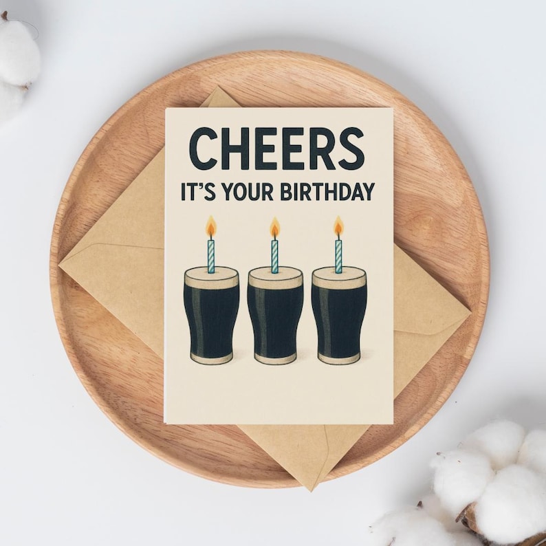 K&ouml;nnte beinhalten: Geburtstagskarte mit dem Text "CHEERS IT'S YOUR BIRTHDAY". Die Karte zeigt drei Cartoon-Biergl&auml;ser mit brennenden Kerzen. Die Karte liegt auf einem Holztablett mit einem braunen Umschlag.