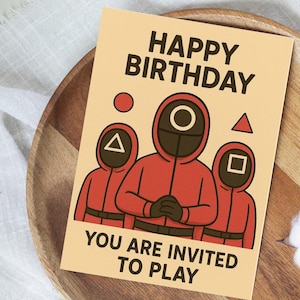 May include: A birthday card with the text "HAPPY BIRTHDAY" at the top and "YOU ARE INVITED TO PLAY" at the bottom. The card features three red-suited figures with different shapes on their masks: a circle, a triangle, and a square.