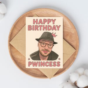 May include: A birthday card with the words "HAPPY BIRTHDAY PWINCESS" in pink lettering. The card features an illustration of a person wearing a hat and glasses, with a crown. The card is on a wooden tray with a brown envelope.