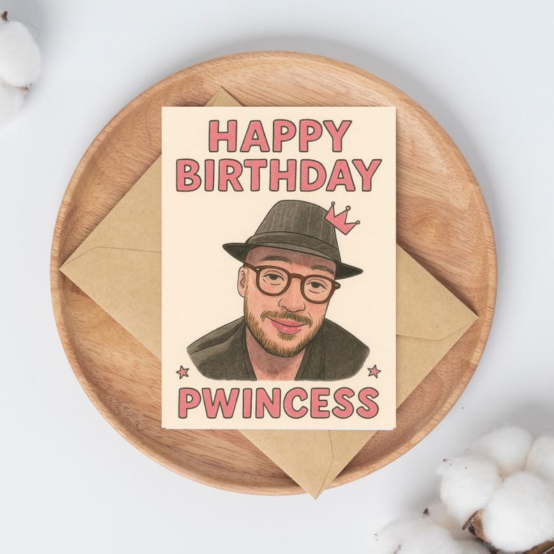 May include: A birthday card with the words "HAPPY BIRTHDAY PWINCESS" in pink lettering. The card features an illustration of a person wearing a hat and glasses, with a crown. The card is on a wooden tray with a brown envelope.