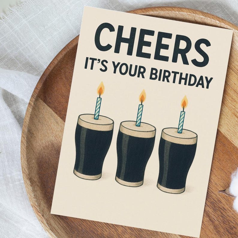 K&ouml;nnte beinhalten: Geburtstagskarte mit dem Text "CHEERS IT'S YOUR BIRTHDAY". Die Karte zeigt drei Cartoon-Biergl&auml;ser mit brennenden Kerzen. Die Karte befindet sich auf einem beigefarbenen Hintergrund und liegt auf einem Holztablett.