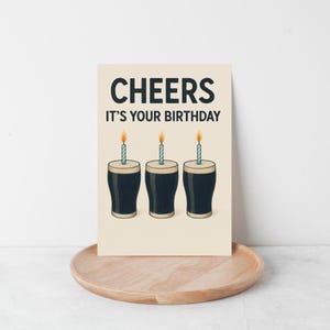 K&ouml;nnte beinhalten: Eine Geburtstagskarte mit dem Text "CHEERS IT'S YOUR BIRTHDAY" in Dunkelblau. Die Karte zeigt drei Cartoon-Pints Stout mit brennenden Kerzen auf cremefarbenem Hintergrund. Die Karte liegt auf einem Holztablett, passend f&uuml;r eine Geburtstagsfeier.