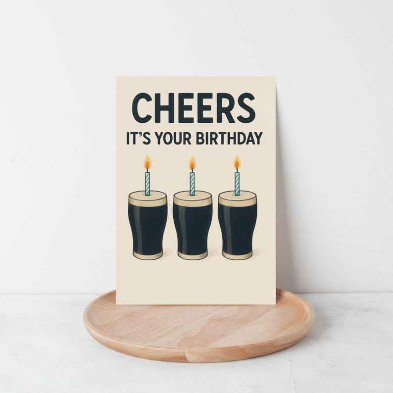 K&ouml;nnte beinhalten: Eine Geburtstagskarte mit dem Text "CHEERS IT'S YOUR BIRTHDAY" in Dunkelblau. Die Karte zeigt drei Cartoon-Pints Stout mit brennenden Kerzen auf cremefarbenem Hintergrund. Die Karte liegt auf einem Holztablett, passend f&uuml;r eine Geburtstagsfeier.