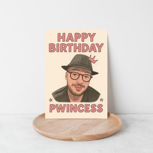 May include: A birthday card with a portrait of a man wearing glasses and a hat, with the words "HAPPY BIRTHDAY" and "PWINCESS" in pink letters. The card is on a wooden tray.