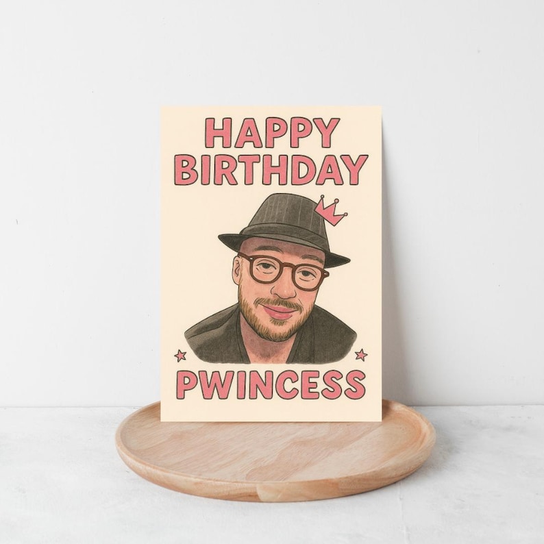 May include: A birthday card with a portrait of a man wearing glasses and a hat, with the words "HAPPY BIRTHDAY" and "PWINCESS" in pink letters. The card is on a wooden tray.