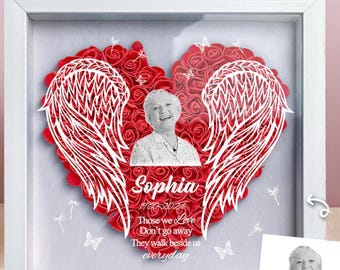 May include: A white frame with a red heart made of paper roses and white angel wings. The heart has a photo of a woman inside. The text on the frame reads "Sophia 1943-2021 Those we love don't go away They walk beside us everyday".