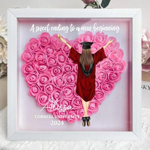 May include: A white shadow box with a pink heart-shaped background made of artificial roses. A graduate in a red cap and gown stands in the center of the heart. The text "A sweet ending to a new beginning" is at the top of the box. The text "Alyssa Cornell University 2024" is at the bottom of the box.