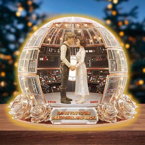 Puede incluir: Un globo de nieve decorativo con una pareja tomados de la mano dentro de una cúpula transparente con un interior de nave espacial. La base tiene rosas doradas y el texto "I Love You to the Death Star and Back". Los nombres "Kevin" y "Emma" también son visibles.