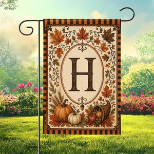 Peut inclure: Un drapeau de jardin rectangulaire présentant un cadre ovale central avec la lettre H dans une police serif foncée. Le motif comprend des feuilles d'automne orange et brunes, des citrouilles, des épis de blé et une corne d'abondance.