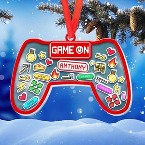 May include: A red-trimmed, controller-shaped Christmas ornament with the text "GAME ON" and "ANTHONY". The ornament features pixelated game icons, including hearts, keys, and flames, and hangs from a red ribbon. The background is a snowy scene.