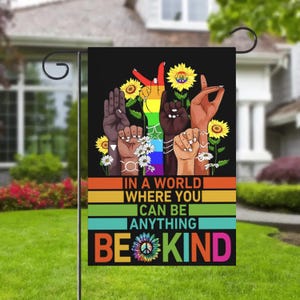 May include: A black garden flag with the message "IN A WORLD WHERE YOU CAN BE ANYTHING BE KIND." The flag features hands in various skin tones, sunflowers, daisies, and a rainbow peace sign.