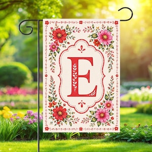 May include: A garden flag with a red letter 'E' in a decorative frame. The flag is cream-coloured with a floral border of red and pink flowers and green leaves. The flag is displayed on a black metal stand.