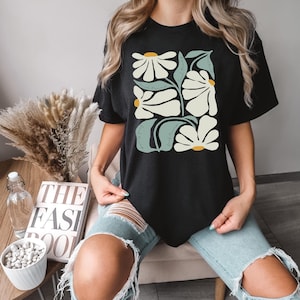 May include: Black t-shirt with a graphic of stylized white daisies and green leaves. The text "feel the love" and "feel the light" is written on the leaves.