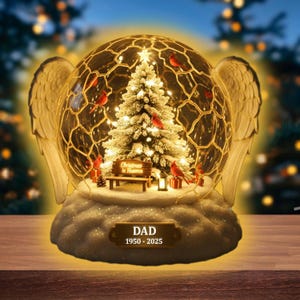Peut inclure: Un globe de neige décoratif sur le thème de Noël. À l'intérieur, un sapin de Noël illuminé est entouré d'oiseaux rouges. Le globe repose sur une base en forme de nuage avec des ailes d'ange. Le texte "DAD 1950-2025" est affiché.