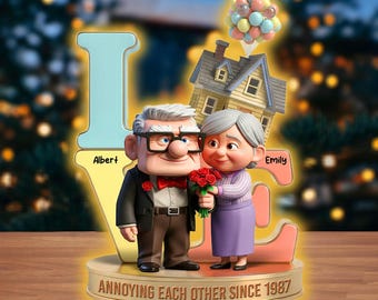May include: A decorative figurine featuring an elderly couple, a house, and balloons, with the word "LOVE" spelled out. The base reads "Annoying Each Other Since 1987." The man wears glasses and a bow tie, while the woman holds roses.