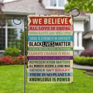 May include: A rectangular garden flag with a distressed, vintage look. The flag features a series of statements in various colors, including "WE BELIEVE," "BLACK LIVES MATTER," and "KNOWLEDGE IS POWER." The flag is hanging on a black metal stand.