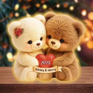 Puede incluir: Dos osos de peluche, uno crema y otro marrón, se abrazan, sosteniendo un corazón rojo. El corazón muestra el año "2025" y los nombres "Ashley & Henry" en una pancarta. Los osos tienen mejillas rosadas, sobre un fondo borroso.