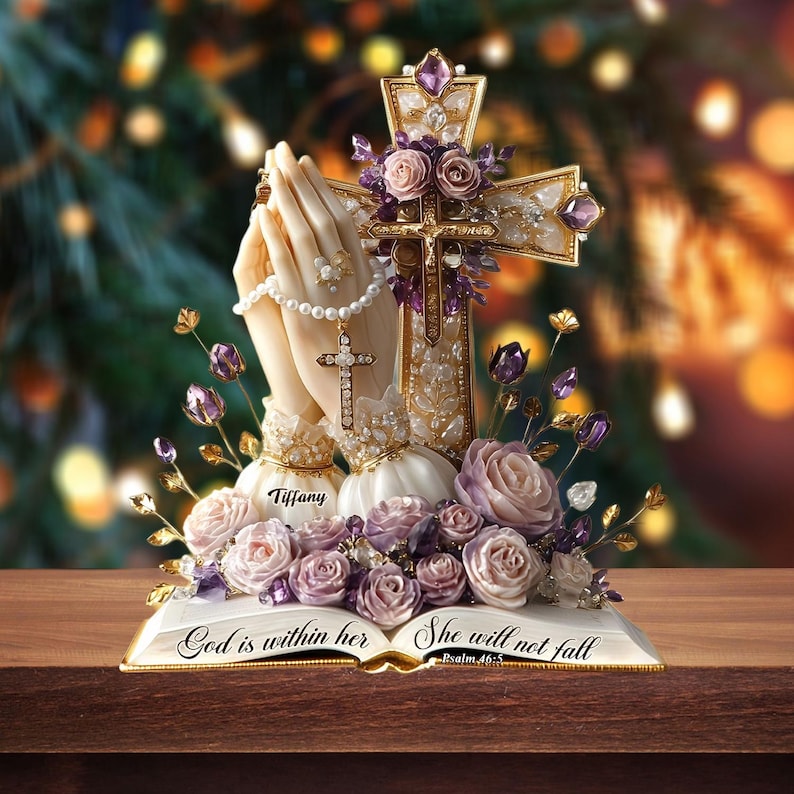 May include: A religious figurine featuring praying hands, a cross, and a book. The hands hold a rosary with a cross pendant. The cross is adorned with purple gems and flowers. The book reads "God is within her, She will not fall".