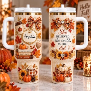 May include: Two cream-coloured tumblers with handles and lids, decorated with 3D-style autumn motifs including orange pumpkins, sunflowers, plaid bows, and fall leaves. One tumbler features the name Sophia, while the other displays the text She believed she could, so she did.