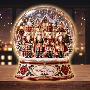 Pode incluir: Um globo de neve de vidro com quatro figuras de quebra-nozes em uniformes vermelhos e dourados sobre uma base nevada. A base é decorada com poinsétias vermelhas e azevinho verde, com o texto The William Family numa moldura dourada.