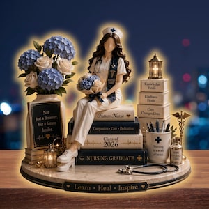 May include: A figurine of a nurse in white scrubs sitting on a stack of books labeled Future Nurse, Class of 2026, and Nursing Graduate. The display includes a bouquet of blue and white flowers, a lantern, a mug, and a caduceus symbol.