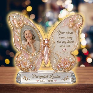 May include: A butterfly-shaped memorial photo frame with a clear crystal base. The frame is pink and gold, with a photo of a woman in one wing and the text "Your wings were ready but my heart was not" on the other. The base reads "Margaret Louise 1950-2026".