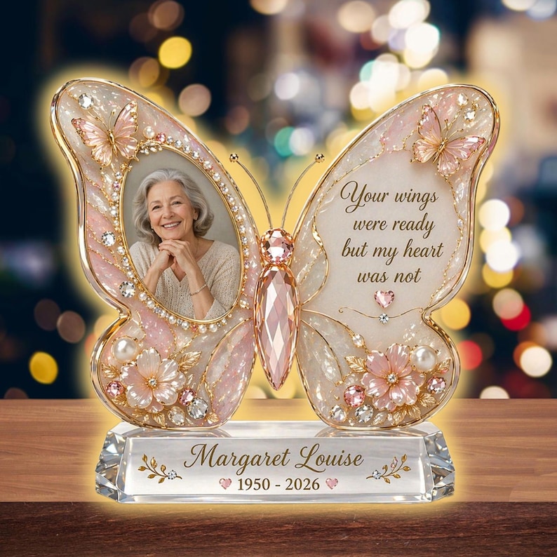 May include: A butterfly-shaped memorial photo frame with a clear crystal base. The frame is pink and gold, with a photo of a woman in one wing and the text "Your wings were ready but my heart was not" on the other. The base reads "Margaret Louise 1950-2026".