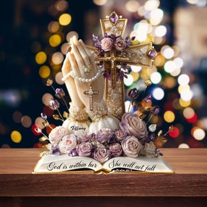 May include: A religious figurine featuring praying hands, a cross, and an open book. The cross is adorned with purple gems and flowers. The book reads "God is within her, she will not fall." The figurine is decorated with pearls and roses.