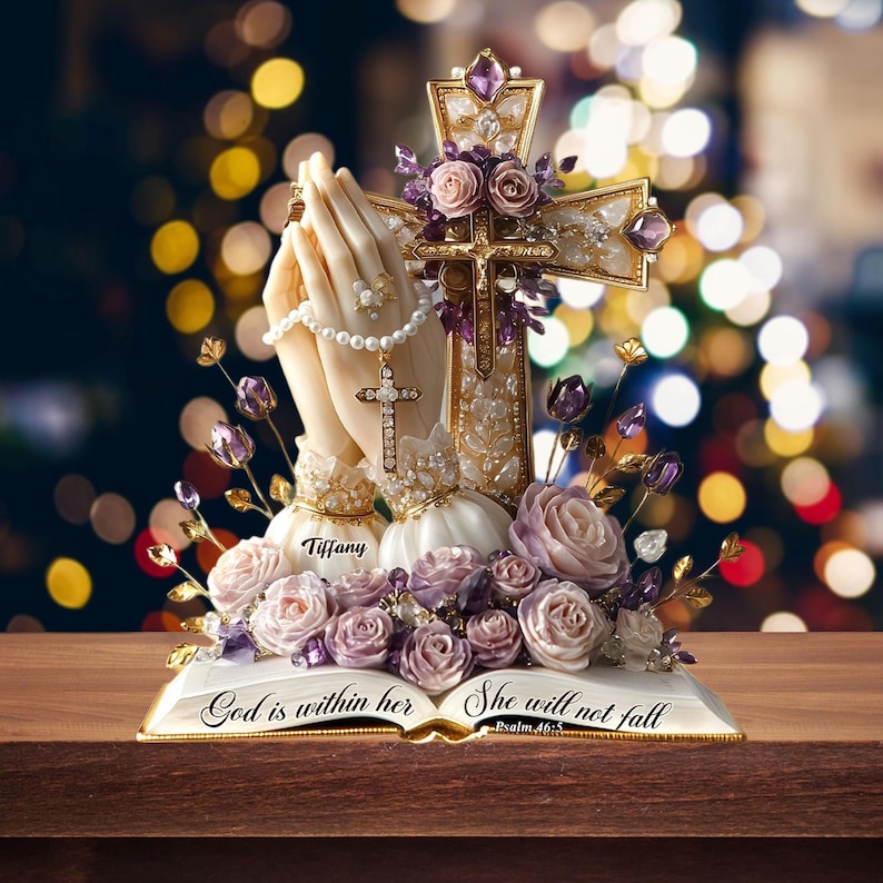 May include: A religious figurine featuring praying hands, a cross, and an open book. The cross is adorned with purple gems and flowers. The book reads "God is within her, she will not fall." The figurine is decorated with pearls and roses.
