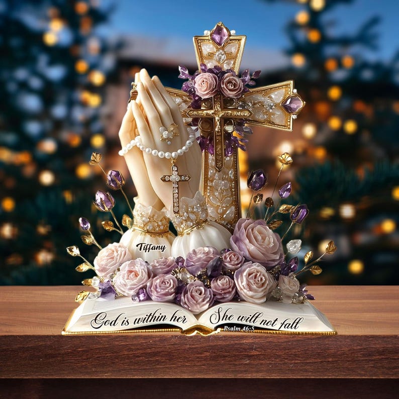 May include: A religious figurine featuring praying hands, a cross, and roses. The cross is adorned with purple gems and gold accents. The base is an open book with the text "God is within her, She will not fall".