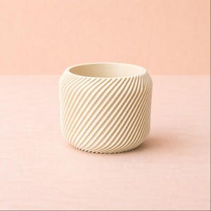May include: A cream-coloured, cylindrical planter with a textured, diagonal ridged pattern. The small, round planter has a wide opening and is suitable for small plants or succulents. The background is a soft pink.