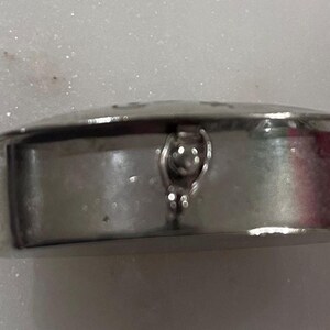 May include: A small, silver-colored, round container with a hinged lid. The container has a decorative clasp and a slightly domed top. The surface appears to be smooth and reflective, suggesting a polished metal finish. The item is likely a trinket box or a similar small storage item.
