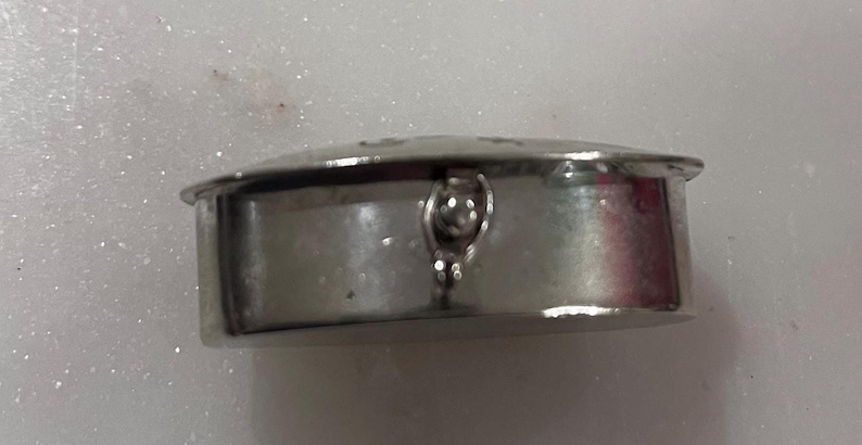 May include: A small, silver-colored, round container with a hinged lid. The container has a decorative clasp and a slightly domed top. The surface appears to be smooth and reflective, suggesting a polished metal finish. The item is likely a trinket box or a similar small storage item.