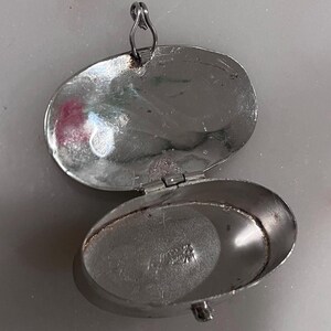 May include: Open silver oval locket with a hinged lid and a small loop for a chain. The locket is open, revealing the interior. The metal has a slightly tarnished appearance. The locket is likely vintage or antique.