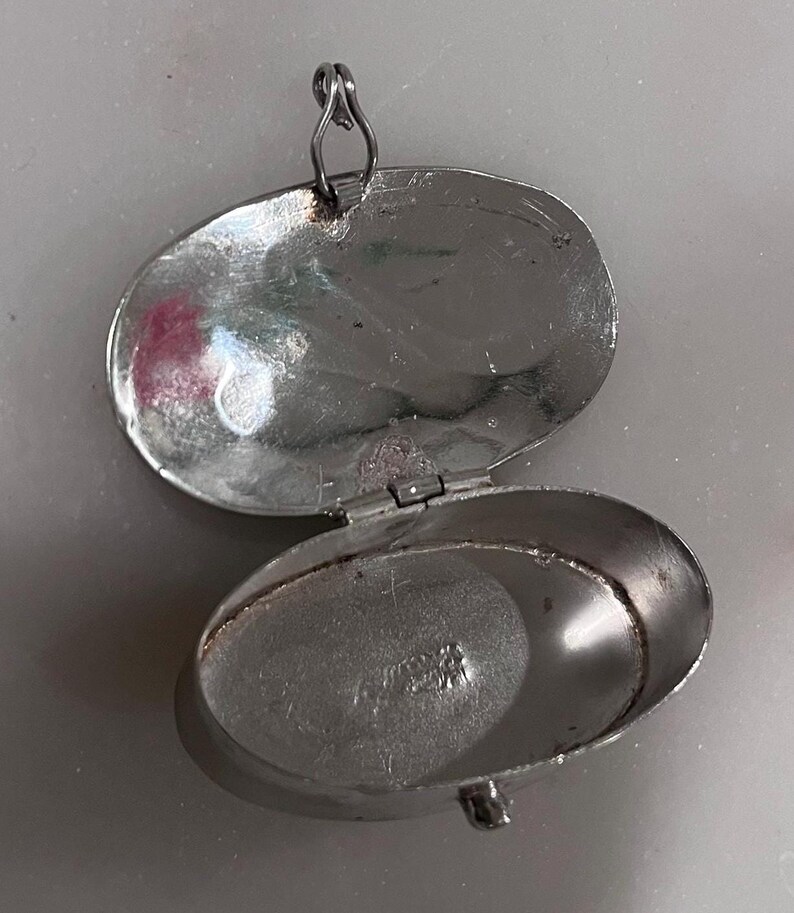 May include: Open silver oval locket with a hinged lid and a small loop for a chain. The locket is open, revealing the interior. The metal has a slightly tarnished appearance. The locket is likely vintage or antique.