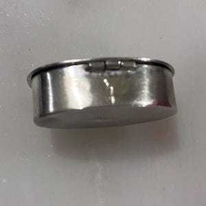May include: A small, oval-shaped, silver-colored metal container with a hinged lid. The container has a slightly hammered texture and a shiny, reflective surface. The lid is closed, and the container appears to be empty.