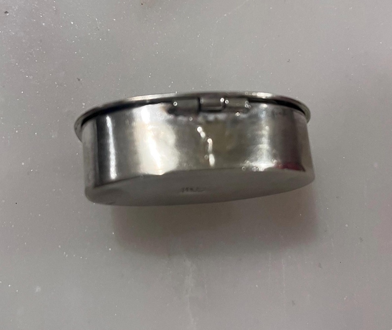 May include: A small, oval-shaped, silver-colored metal container with a hinged lid. The container has a slightly hammered texture and a shiny, reflective surface. The lid is closed, and the container appears to be empty.