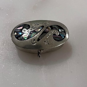 May include: Oval-shaped silver trinket box with an abalone inlay design. The lid features cut-out patterns and is hinged. The abalone inlay displays iridescent colors of blue, green, and purple. The box is resting on a white surface.