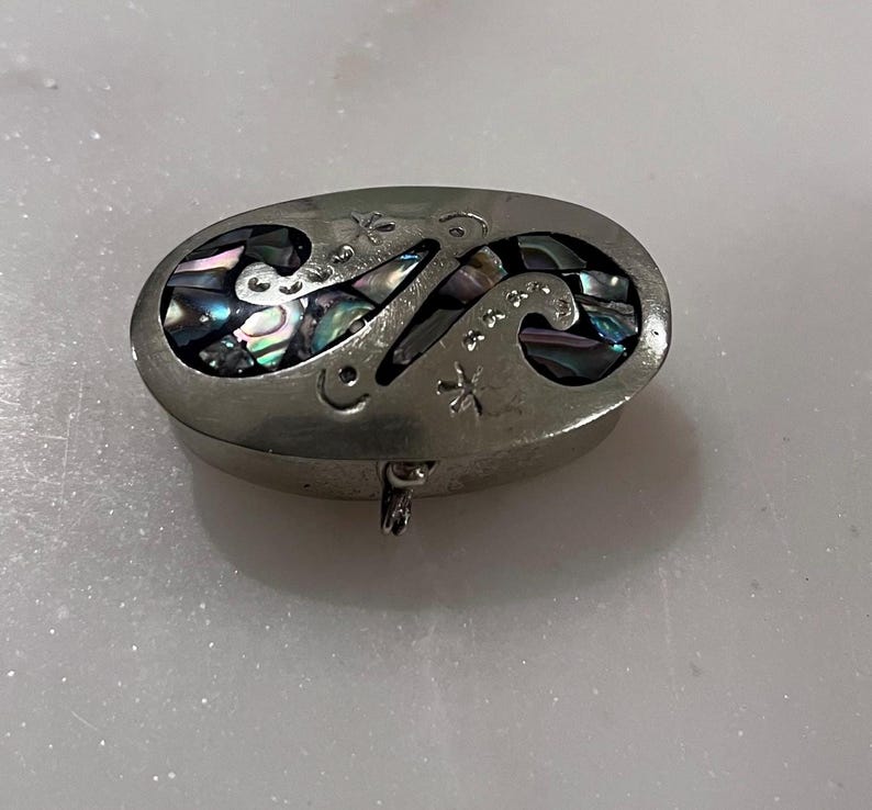 May include: Oval-shaped silver trinket box with an abalone inlay design. The lid features cut-out patterns and is hinged. The abalone inlay displays iridescent colors of blue, green, and purple. The box is resting on a white surface.